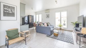 Un salon spacieux avec un canapé gris, un petit coin repas, une cuisine équipée d’un réfrigérateur et un téléviseur.