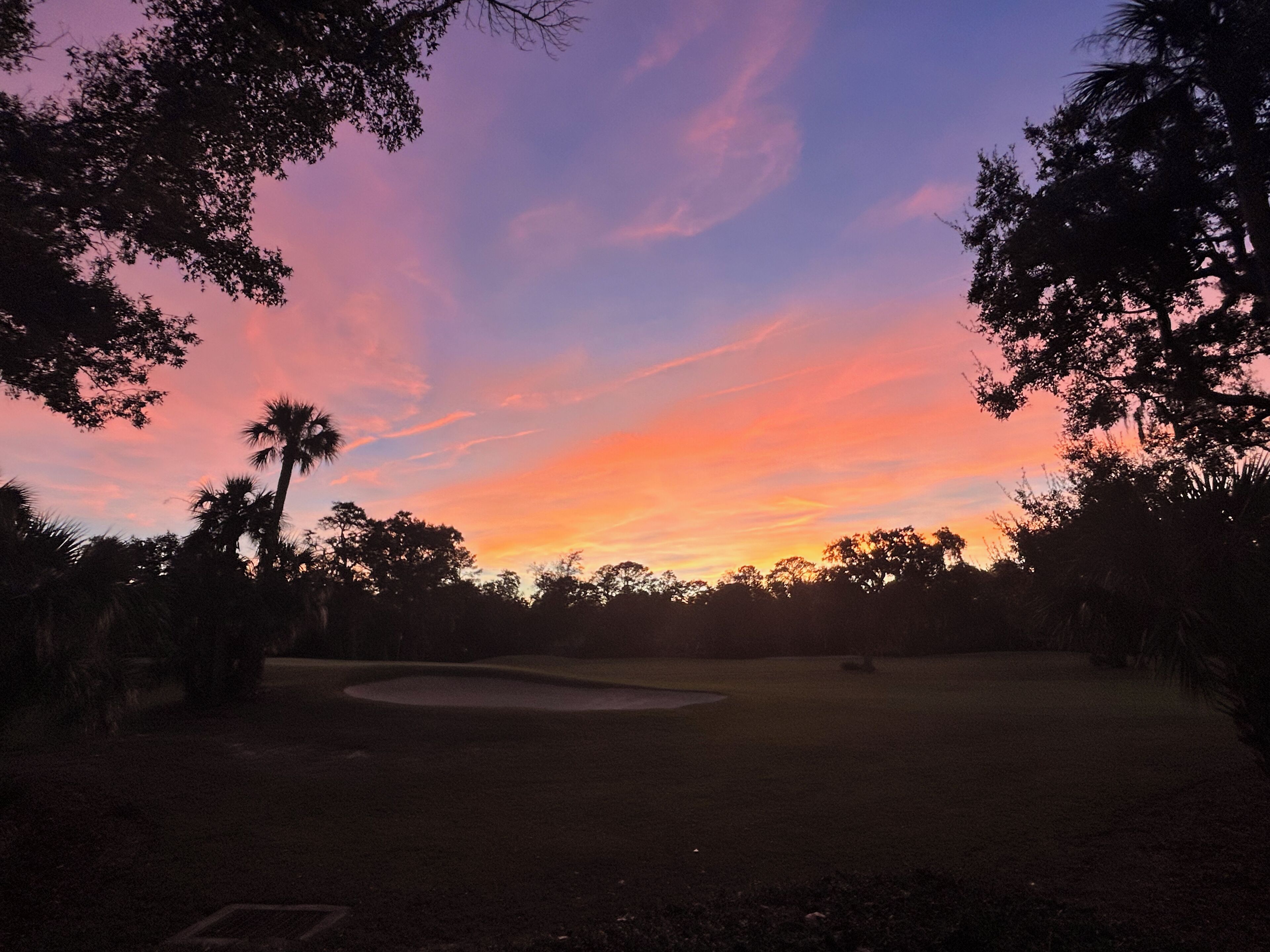 A golf course at sunset with trees and a sand bunker.