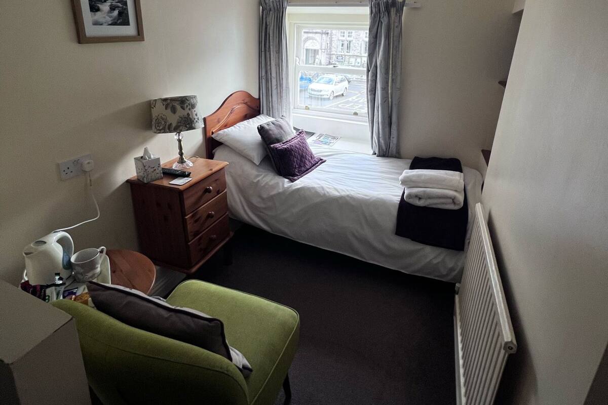 Ein kleines Hotelzimmer mit einem Bett, einem Schreibtisch, einem Stuhl, einem Heizkörper, einem Fenster mit Vorhängen und einem Bild an der Wand.