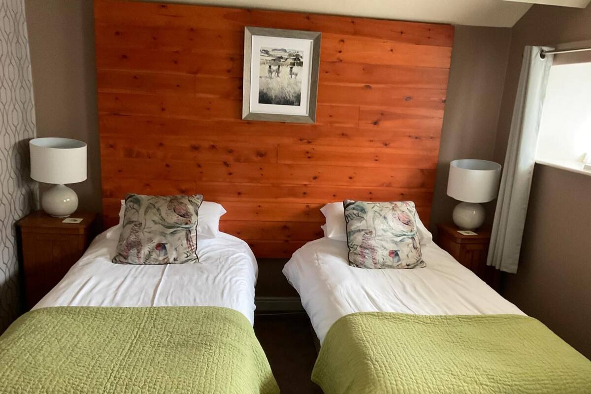Ein Zimmer mit zwei Betten, jeweils mit grüner Bettdecke und floralen Kissen, einem Holzkopfende und einem gerahmten Bild darüber.