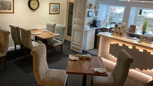 A dining area with a wooden table and chairs, a clock on the wall, and a kitchen area visible through an open door.