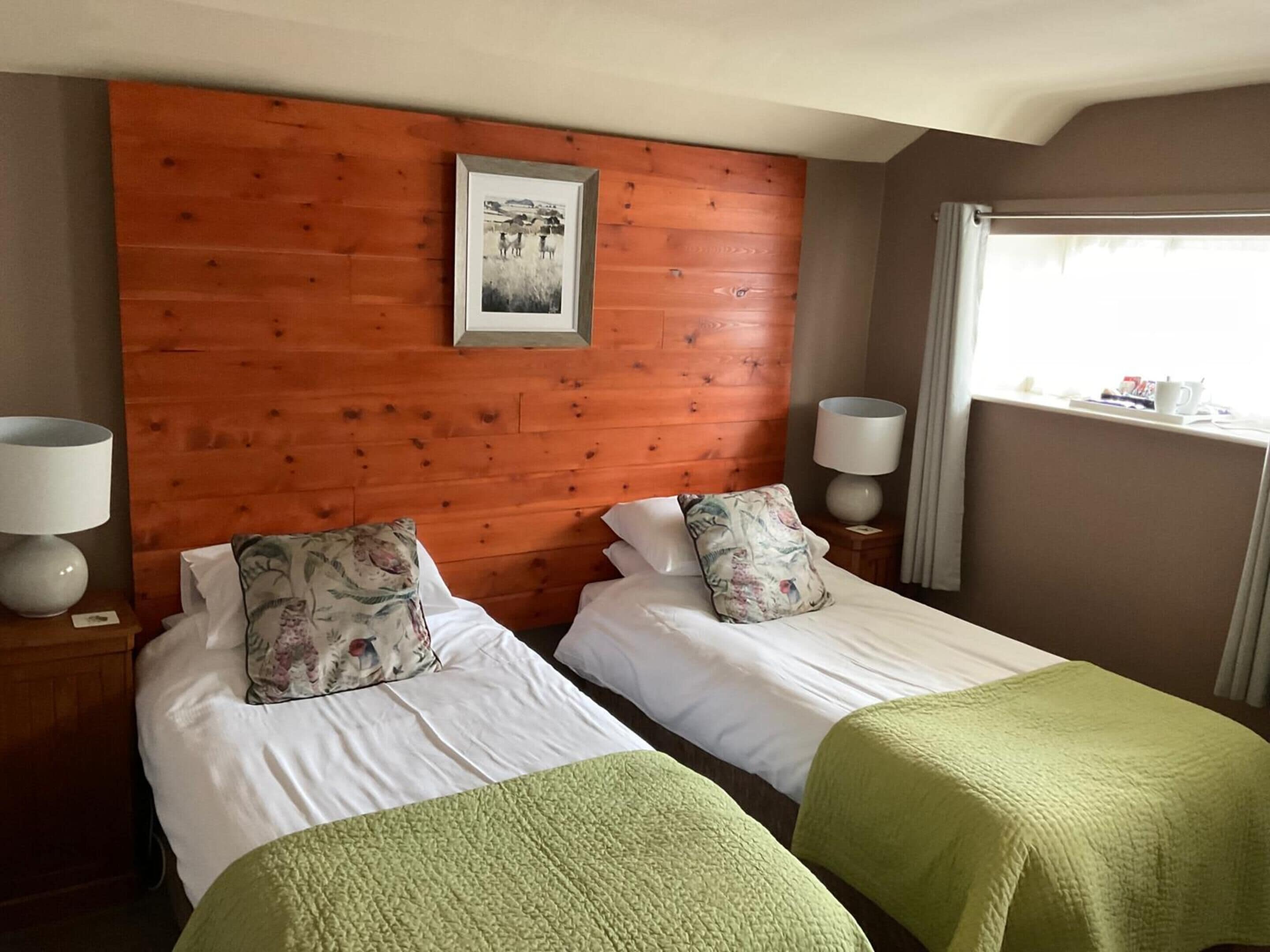 Camera d'albergo con due letti singoli, una testiera in legno, un quadro incorniciato, lampade da comodino e una finestra con tende.