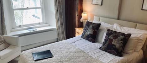 Un dormitorio ordenado con cama, una mesita de noche con lámpara, una ventana con cortinas y un cuadro enmarcado en la pared.