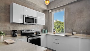 A modern kitchen with white cabinets, a stainless steel microwave, a stove, and a large window offering a view of greenery and buildings. - Boho Loft | Modern & Chic in Santurce (San Juan)