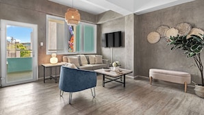 A modern living room with a sofa, blue armchair, coffee table, and a bench. A TV is mounted on the wall, and there is a large window with a view of buildings. - Boho Loft | Modern & Chic in Santurce (San Juan)