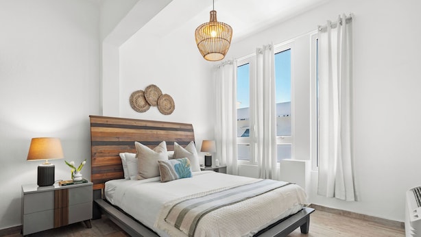 A modern bedroom with a wooden headboard, a large bed, a bedside table with a lamp, and a view of the outdoors through a window. - Boho Loft | Modern & Chic in Santurce (San Juan)