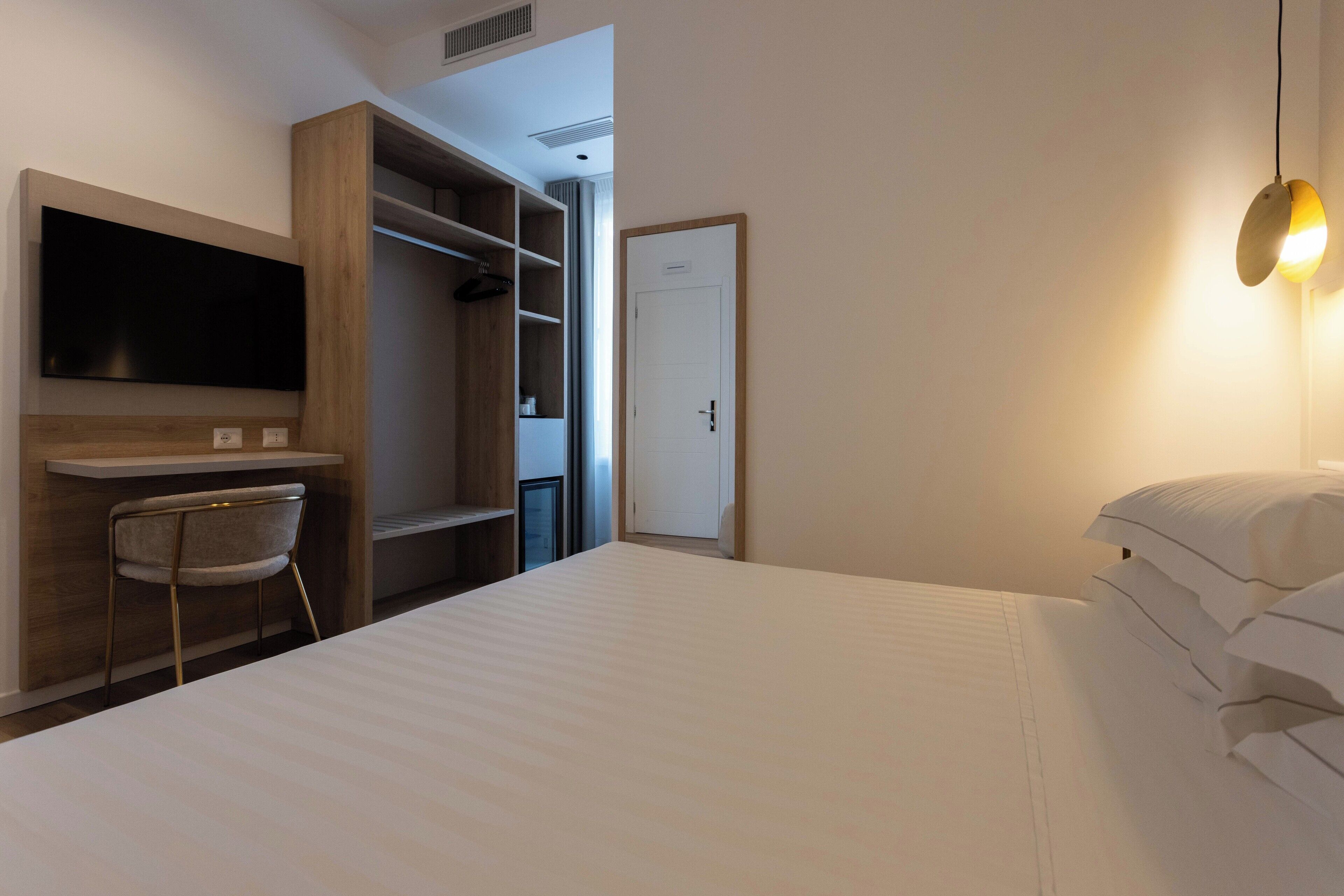 Quarto de hotel com uma cama, uma mesa com cadeira, televisão na parede e um guarda-roupa.
