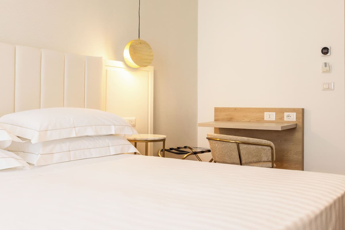 A hotel room with a bed, a bedside table, a chair, a desk, and a wall-mounted shelf.