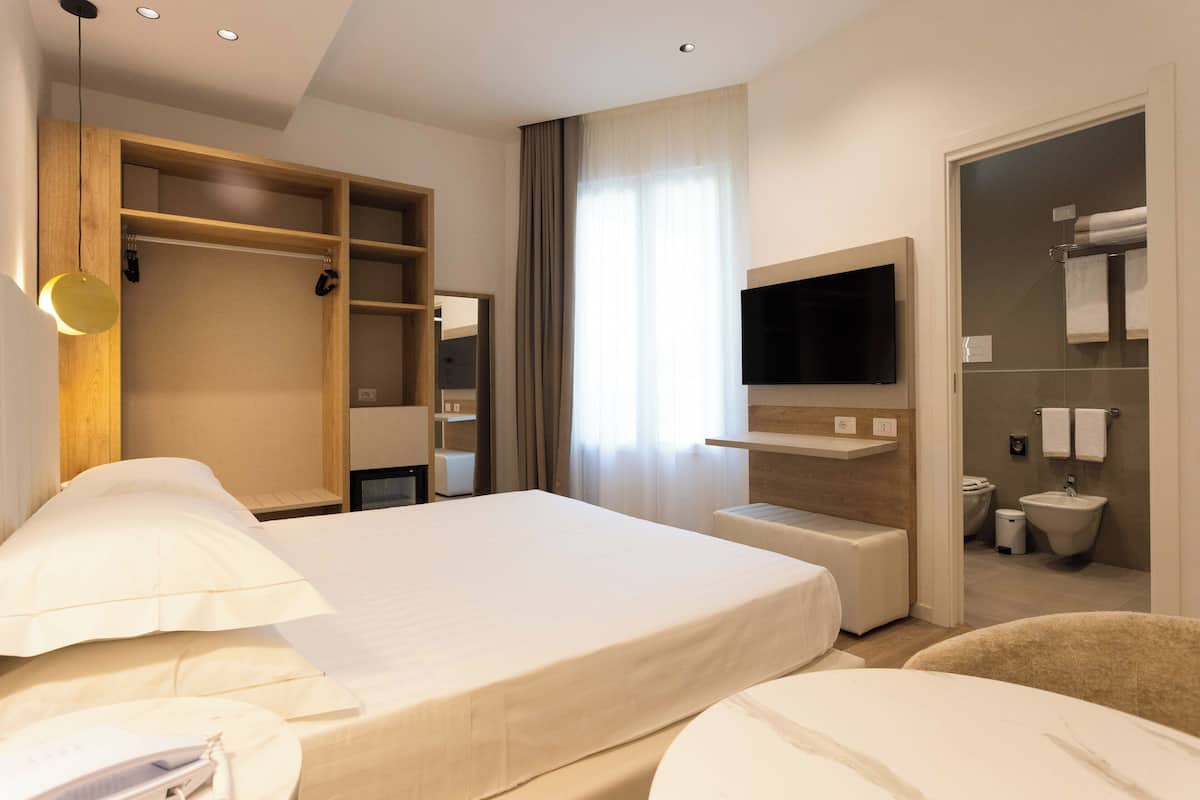 A modern hotel room with a large bed, a wooden wardrobe, a television, and a bathroom with a sink and toilet.
