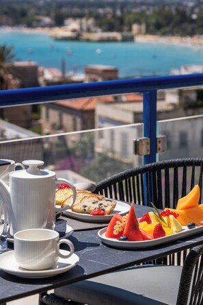 Mesa en balcón con cafetera, taza y plato de fruta y pasteles, con vista a una playa y edificios.