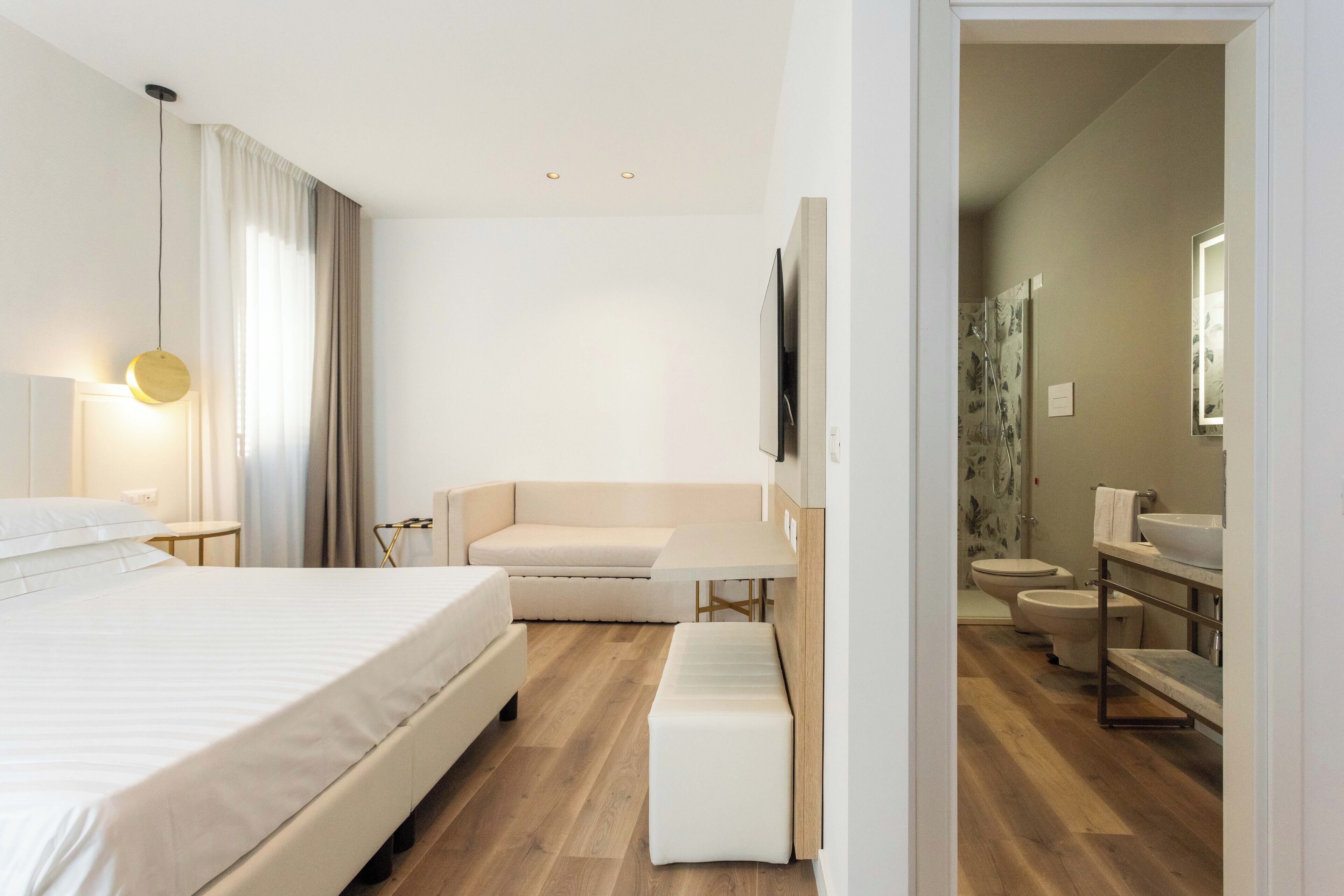 A modern hotel room with a large bed, a sofa, a small table, and a bench. The room has wooden flooring and a large mirror on the wall.