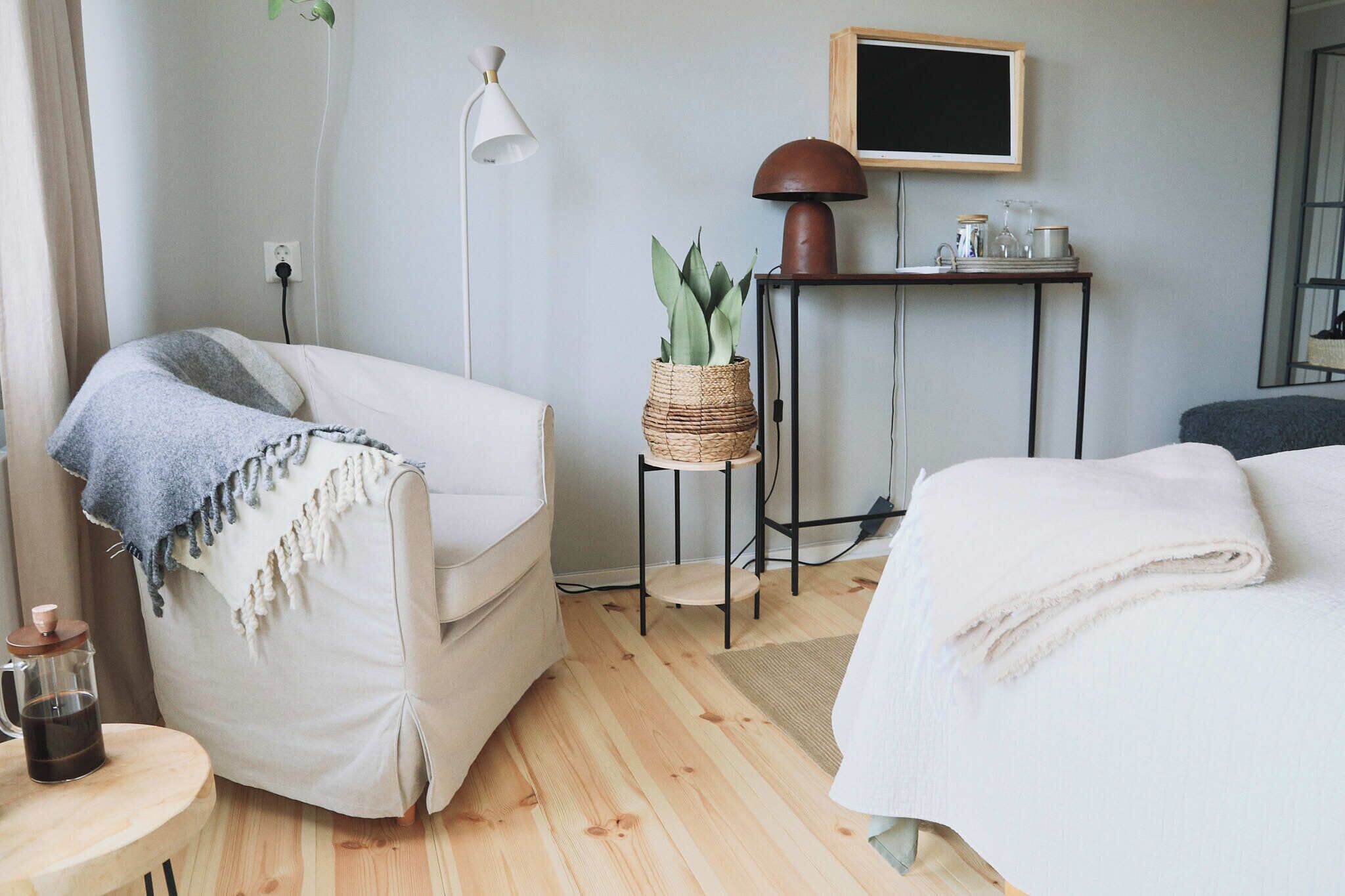 Una habitación de hotel acogedora con suelo de madera, un sillón blanco con una manta azul, una mesita redonda con una cafetera, una mesita de noche con una lámpara y una cama con un edredón blanco.