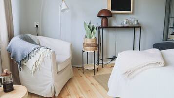 Una habitación de hotel acogedora con suelo de madera, un sillón blanco con una manta azul, una mesita redonda con una cafetera, una mesita de noche con una lámpara y una cama con un edredón blanco.