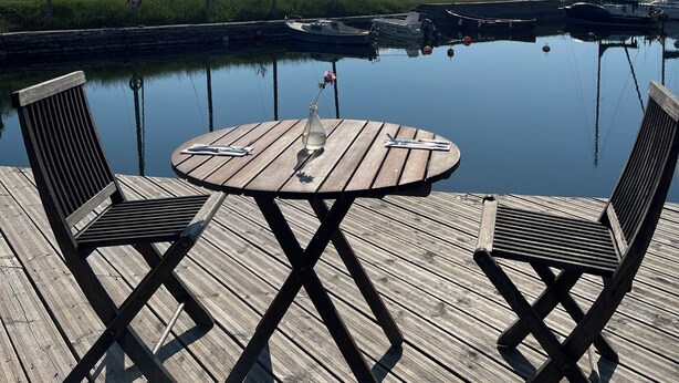 A wooden deck with a round table and two chairs, overlooking a calm body of water with boats and a distant shoreline. - Vallevikens Hotell och Restaurang  (Lärbro)