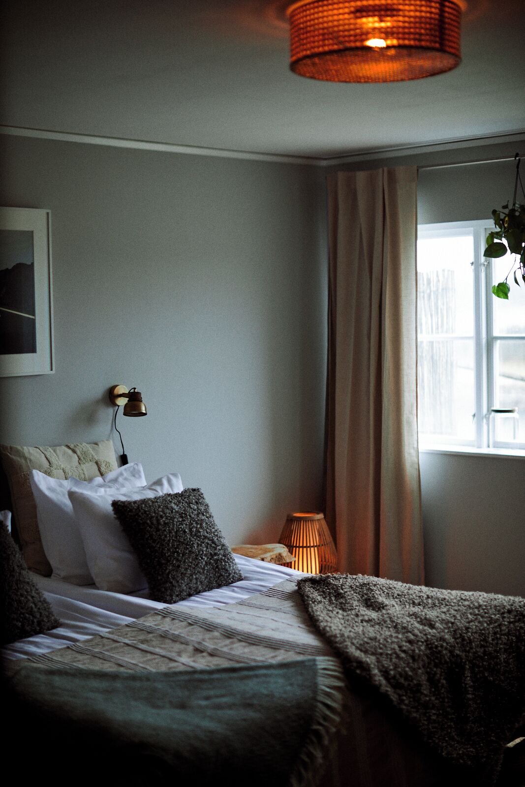 Un dormitorio con cama, lámpara de noche, ventana con cortinas y lámpara de pared.