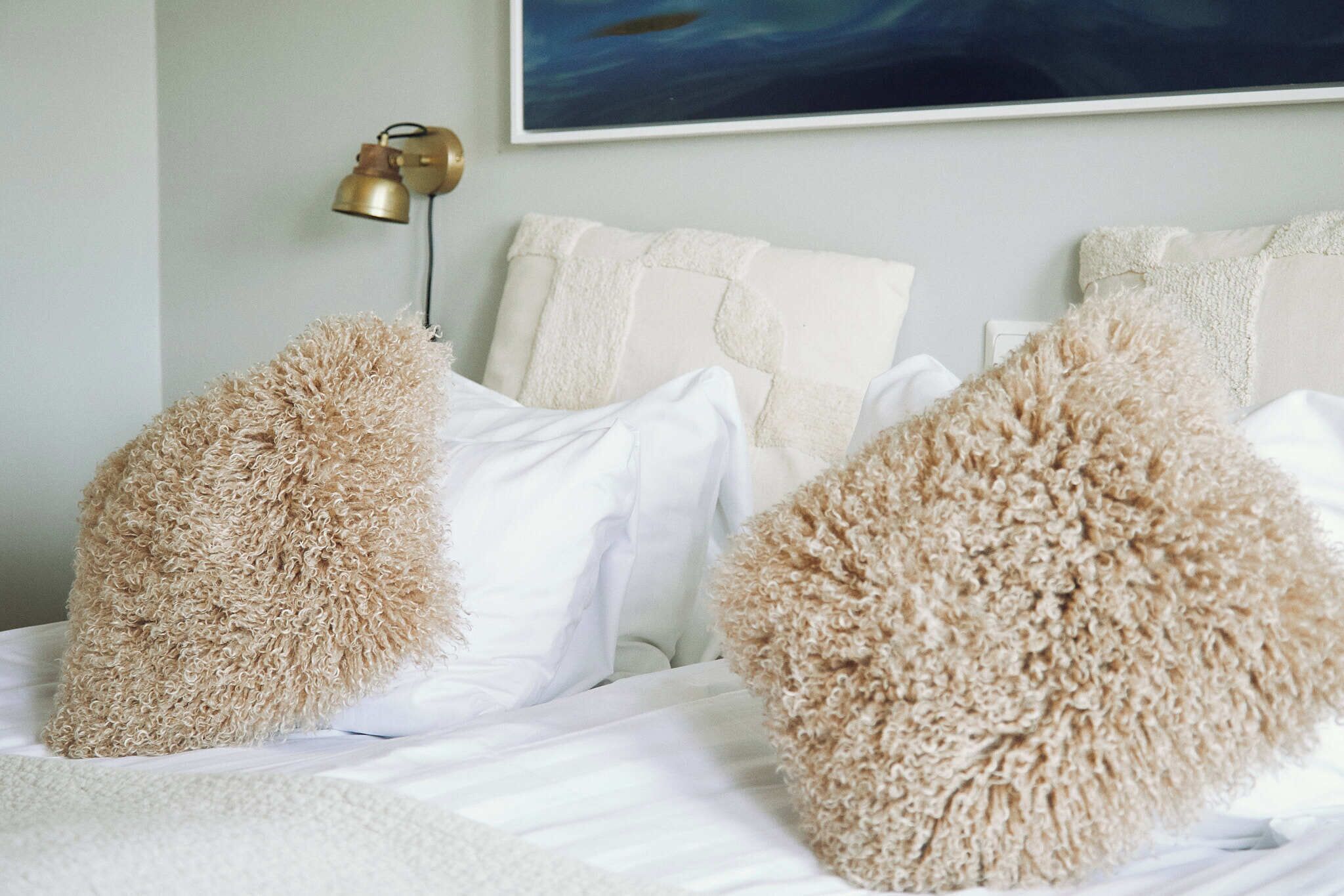Un lit avec du linge de lit blanc et de moelleux oreillers beige, une lampe dorée et un tableau accroché au mur.