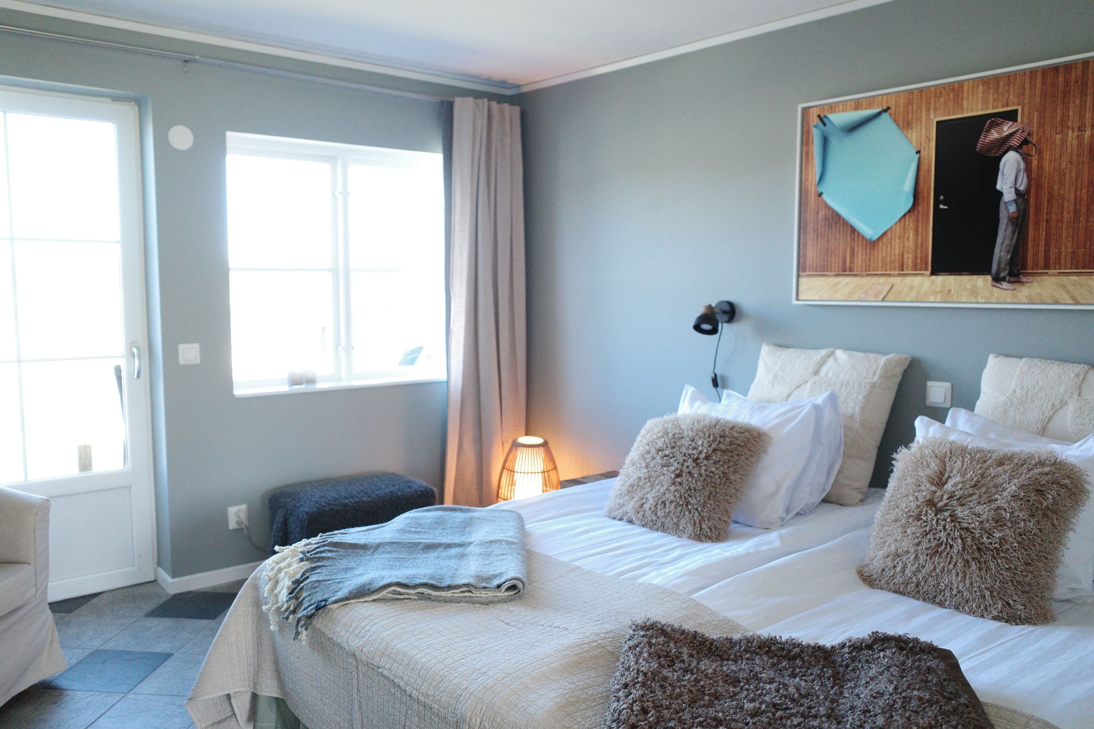 A hotel room with a bed, pillows, a bedside table, a lamp, and a window with curtains.