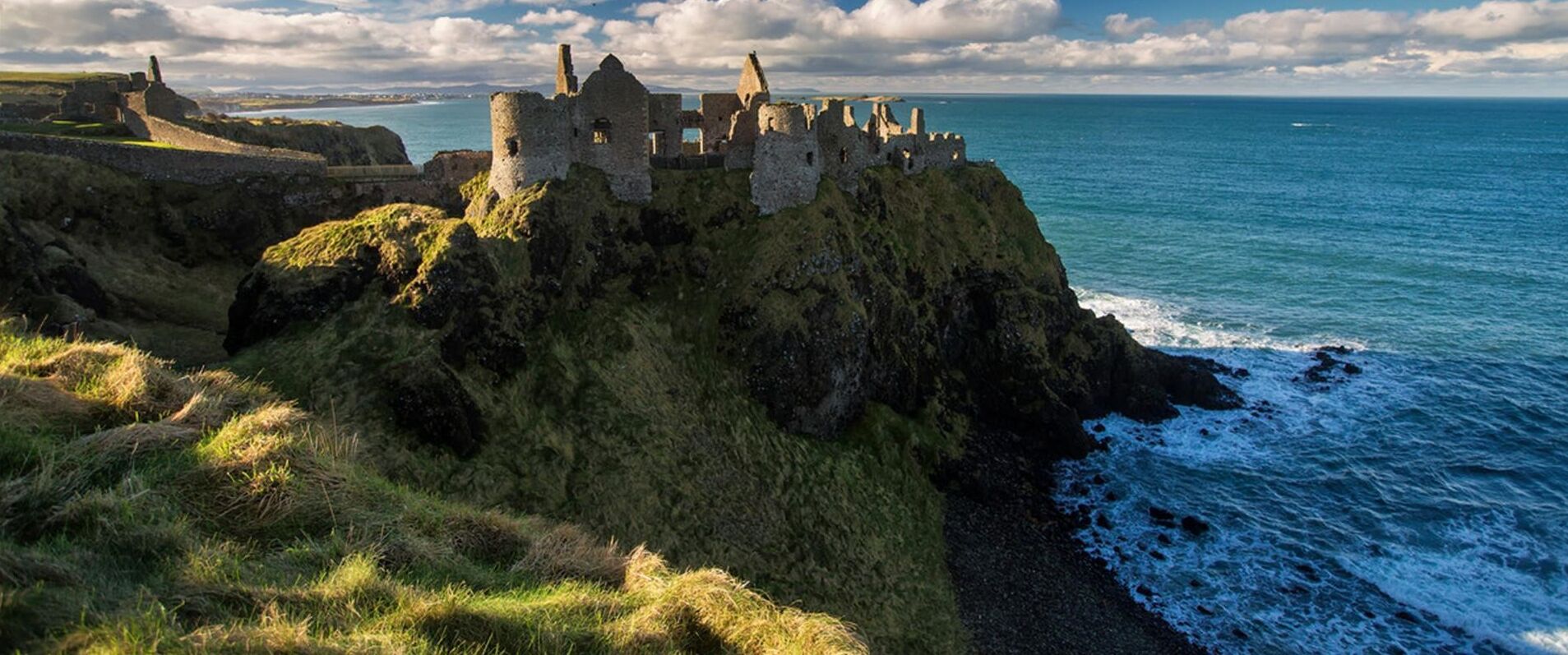 Ruínas de um castelo costeiro com vista para o mar e paisagem distante.
