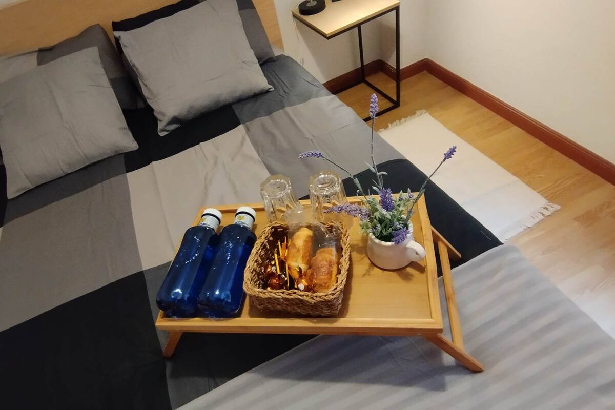 A neatly made bed with a checkered bedspread, a bedside table with a lamp, and a tray with food and drinks on the floor.