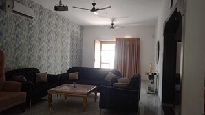 A living room with a sofa, armchair, and coffee table. There is a ceiling fan, a window with curtains, and a statue on a stand. - Fursat Villa (Mohali)