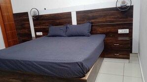 A bed with a dark blue mattress and pillows, a wooden headboard with a ship model, and a white wall with a power outlet. - Fursat Villa (Mohali)