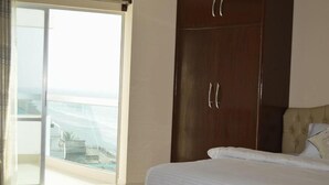 Un dormitorio con cama, armario y puerta corrediza de vidrio que da a un balcón con vista a la playa y edificios.
