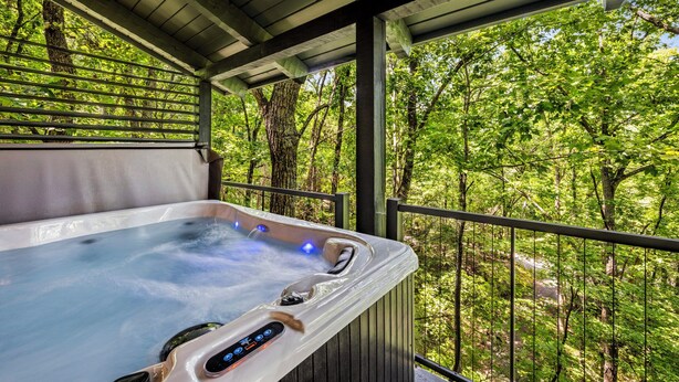 Un jacuzzi con panel de control digital, rodeado por una terraza de madera y una barandilla metálica, con vistas a una zona boscosa.
