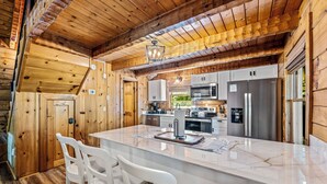 Interior de una cabaña de madera con cocina, que incluye una isla grande, sillas blancas y electrodomésticos modernos.