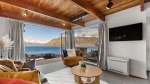Ein modernes Wohnzimmer mit großem Fenster, das Blick auf Berge und See bietet, einem Flachbildfernseher und einer gemütlichen Sitzecke mit Sofa und Sesseln.