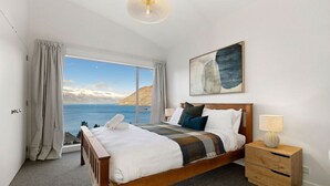 Ein Schlafzimmer mit großem Fenster, das Meer und Hügel zeigt, einem hölzernen Bett, einem Nachttisch mit Lampe sowie einem Gemälde an der Wand.