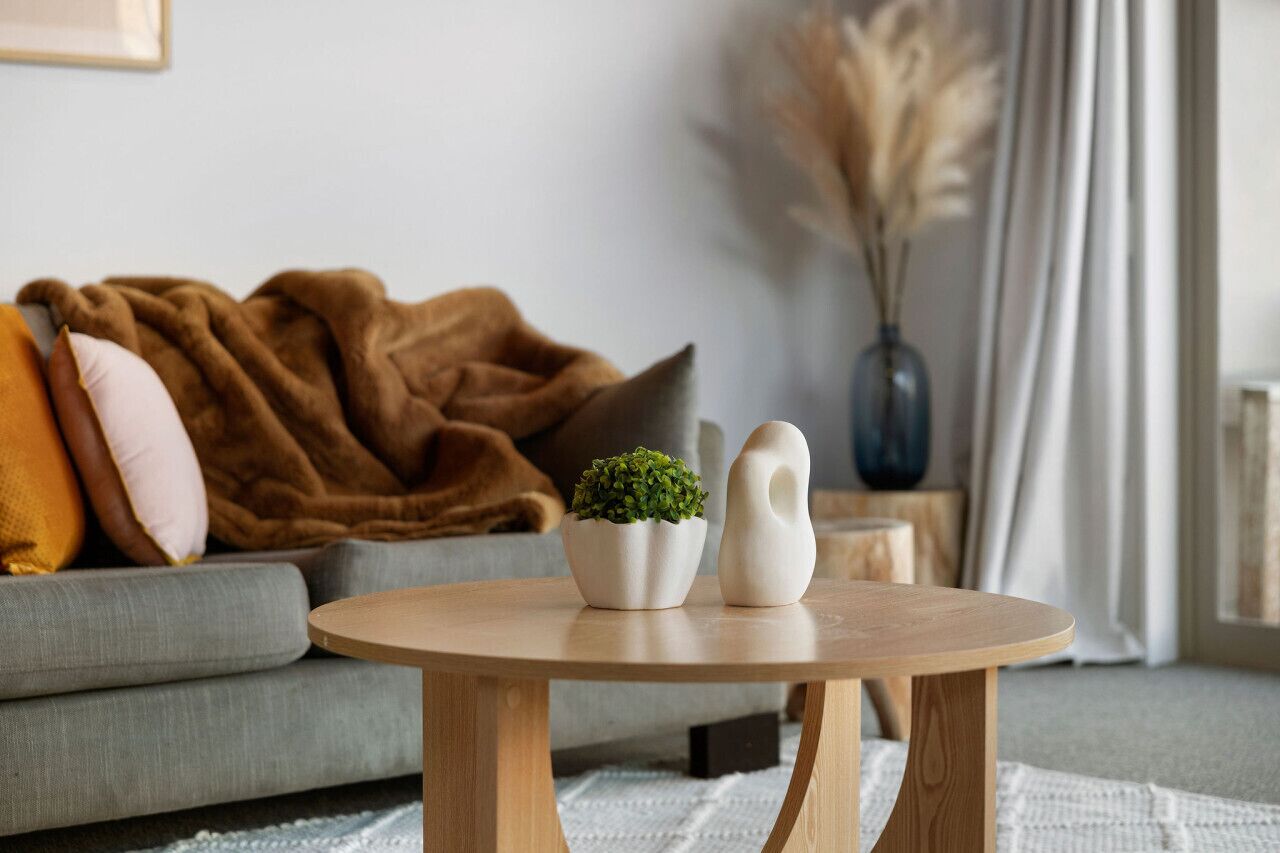 A cozy living room with a grey sofa, a brown blanket, a small wooden table with two white vases and a green plant, and a vase with dried pampas grass.