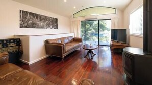 A living room with a sofa, a coffee table, a fireplace, and a large window with a view of trees. - Clematis Hideaway (Halls Gap)