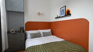 Ein Schlafzimmer mit einem Bett, einem Nachttisch, einem an der Wand befestigten Regal mit einem gerahmten Bild und einer gemusterten Tagesdecke.