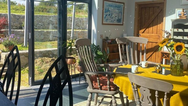 Uma sala de jantar iluminada pelo sol, com uma mesa amarela, cadeiras e vista através de grandes portas de vidro.