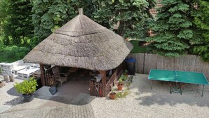 Un espace extérieur aménagé avec une toiture de chaume, comprenant une table et des chaises, entouré de plantes en pot et d’une table de ping-pong.