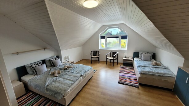 Une chambre avec un plafond en pente, une grande fenêtre, deux lits, une chaise et une petite table.