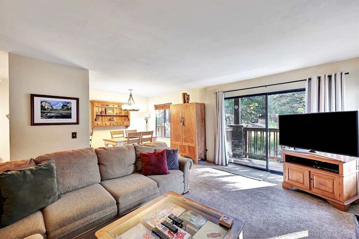 A living room with a sofa, coffee table, and a TV on a wooden stand. There is a dining area with chairs and a table, and a large window with curtains.