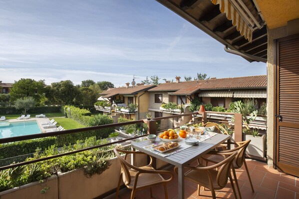 A balcony with a table set for breakfast, overlooking a swimming pool and a garden.