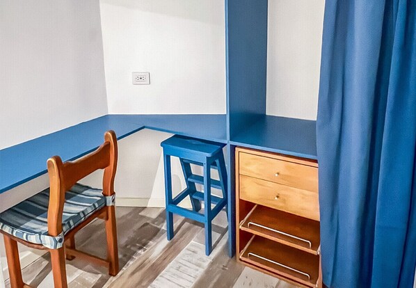 A corner room with a wooden chair, a blue stool, and a wooden desk with drawers.