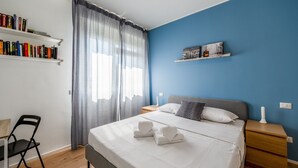 Ein Schlafzimmer mit einem Bett, einem Sessel, einem Schreibtisch, einem Bücherregal und einem Fenster mit Vorhängen.