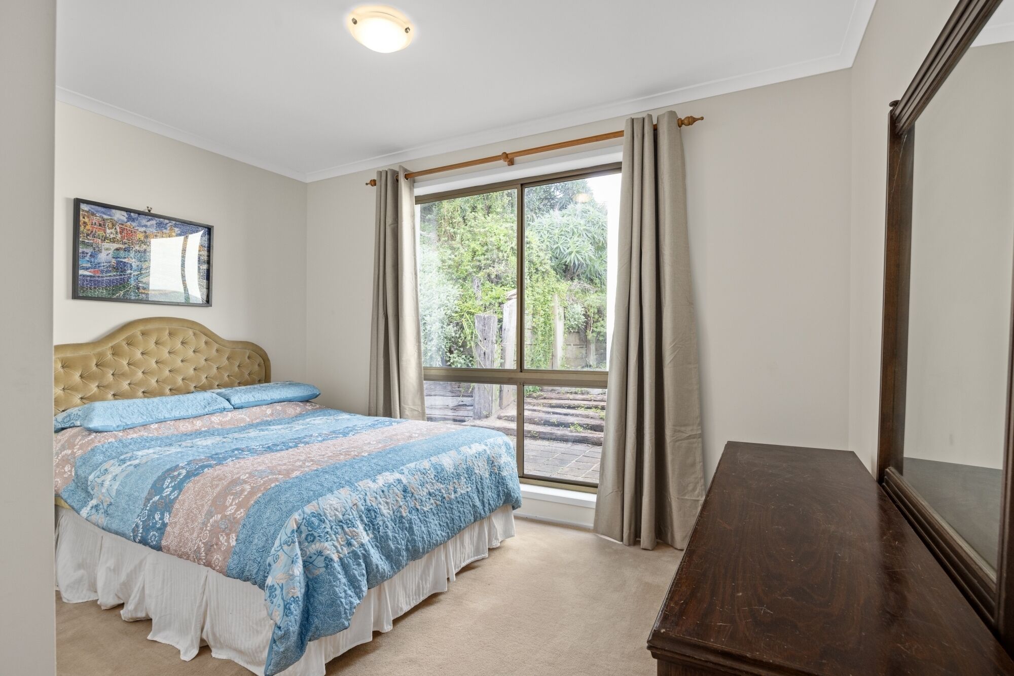 Une chambre à coucher avec un lit, une commode en bois, une fenêtre avec des rideaux et un tableau encadré au mur.