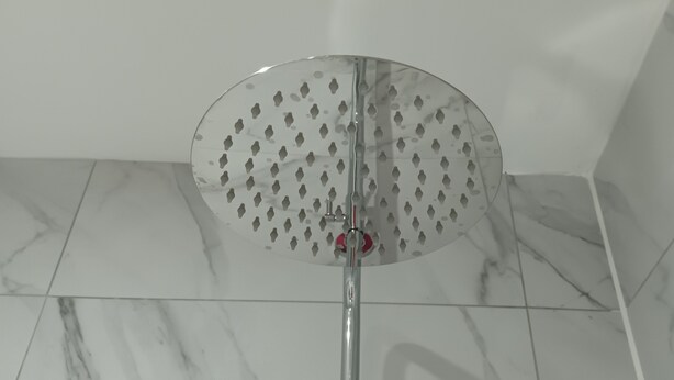 Un cabezal de ducha con diseño circular y perforado, montado sobre un poste metálico, sobre un suelo de baldosas con estampado de mármol.