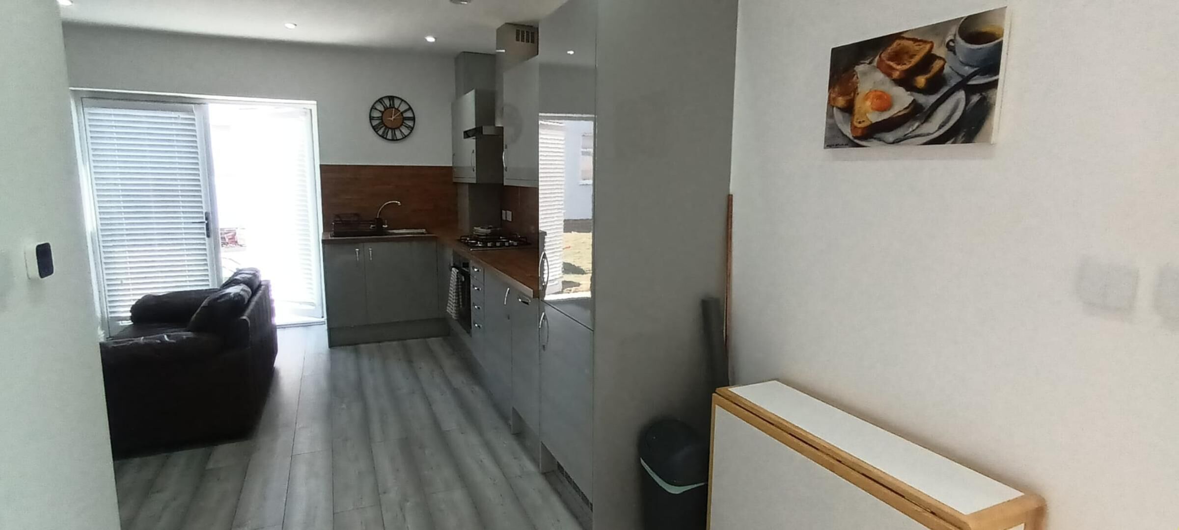 A modern kitchen with a built-in oven, a sink, and a clock on the wall. There is a dining area with a table and chairs visible through the window.