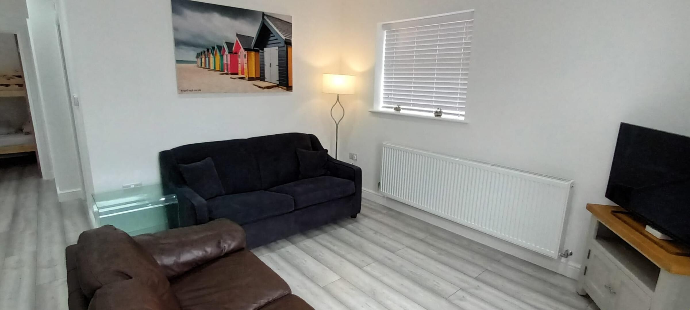 A living room with a dark blue sofa, a brown armchair, a glass coffee table, a white radiator, a TV on a wooden stand, and a painting of colorful beach huts.