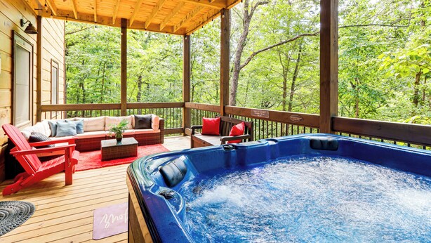 Une terrasse en bois avec un jacuzzi, des chaises rouges et un canapé avec des coussins.