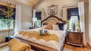 Une chambre à coucher avec un grand lit, une tête de lit en bois, deux tables de chevet avec des lampes, une table de nuit, une fenêtre avec des rideaux et un tableau encadré au mur.