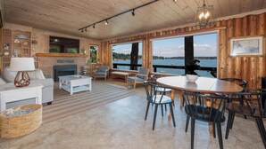 A spacious living room with a fireplace, large windows overlooking a body of water, and a dining area with a round table and chairs. - Waterfront Vashon Island Home, Beach Access & View (Vashon Island)