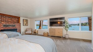 A bedroom with a brick fireplace, a bed, a TV, a view of the water, and a large window. - Waterfront Vashon Island Home, Beach Access & View (Vashon Island)