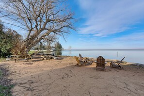 Una spiaggia sabbiosa con un grande albero, tavoli da picnic e poltrone Adirondack a bordo di un lago tranquillo.
