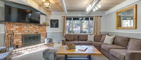 A cozy living room with a brick fireplace, a large flat-screen TV, a brown leather sofa, and a wooden coffee table.